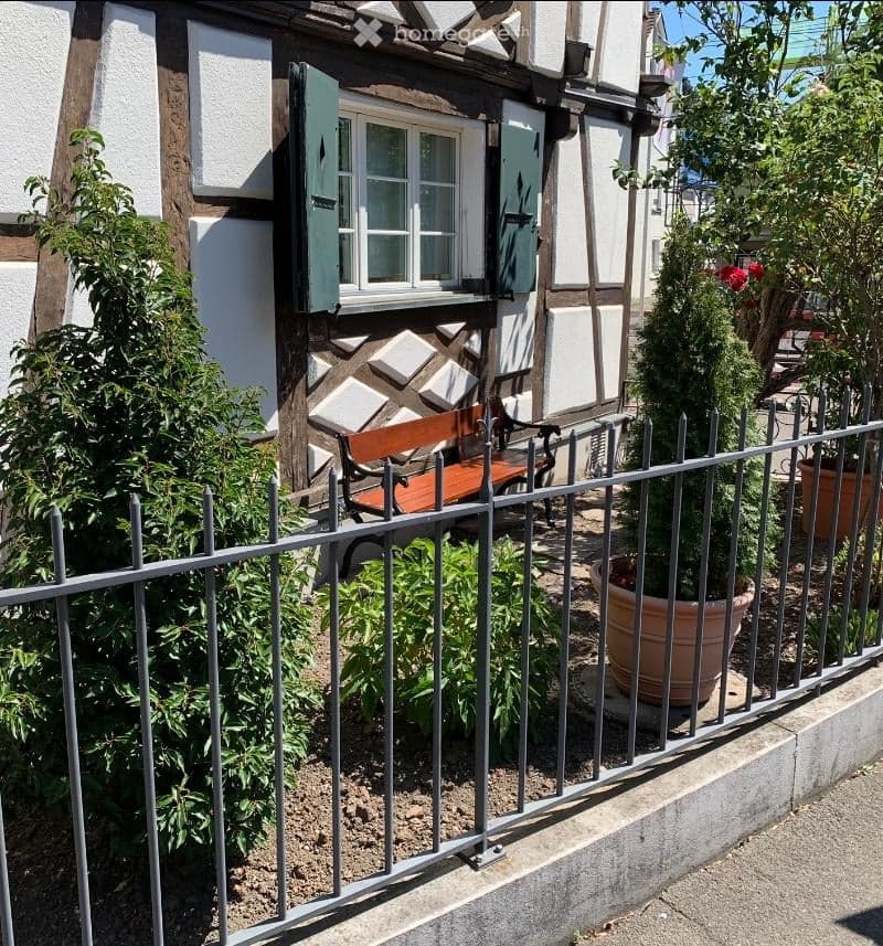 A quaint garden area in front of a traditional half-timbered house, featuring a metal fence, potted plants, and a wooden bench.