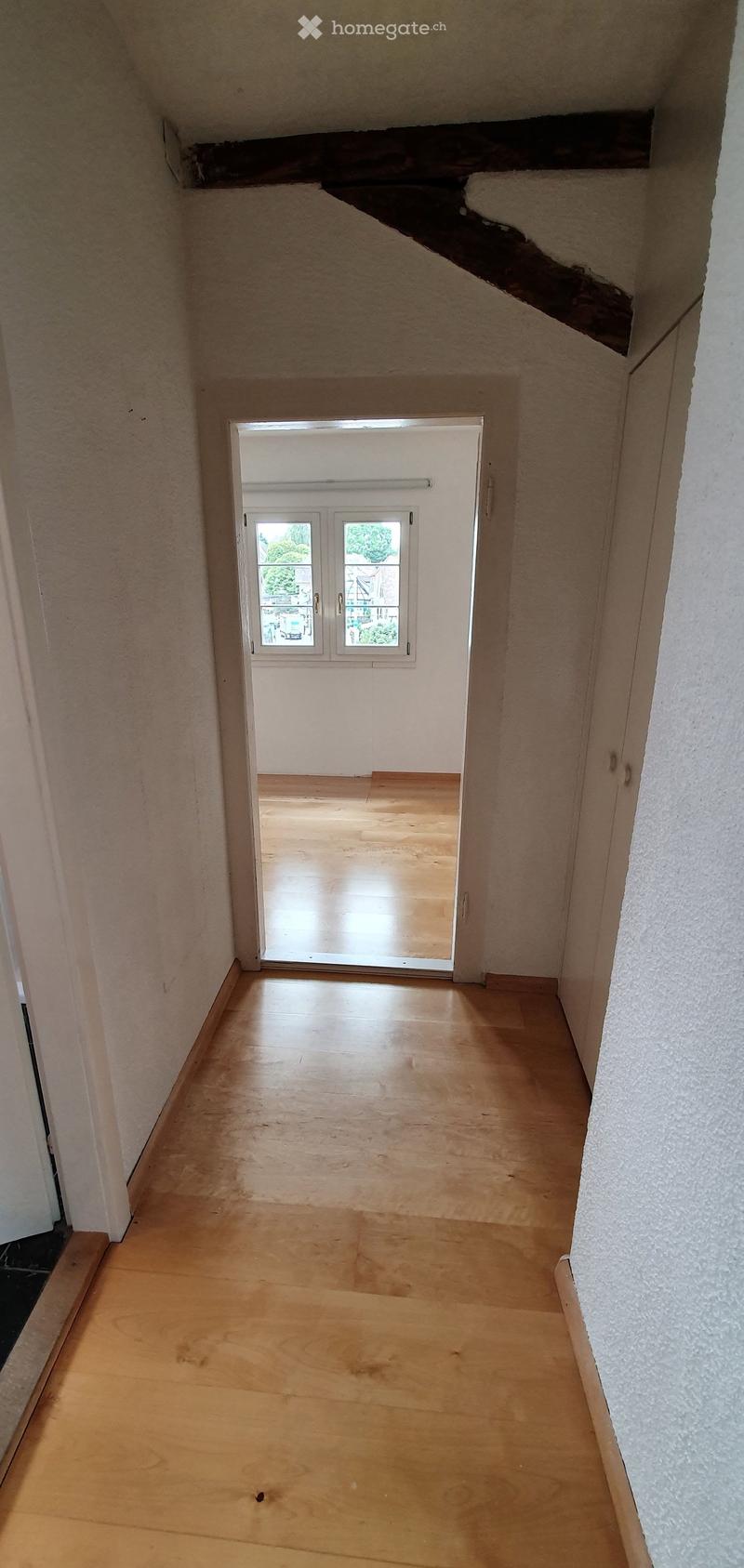 Empty hallway with wooden flooring and white walls, leading to a room with a window and wooden floor.