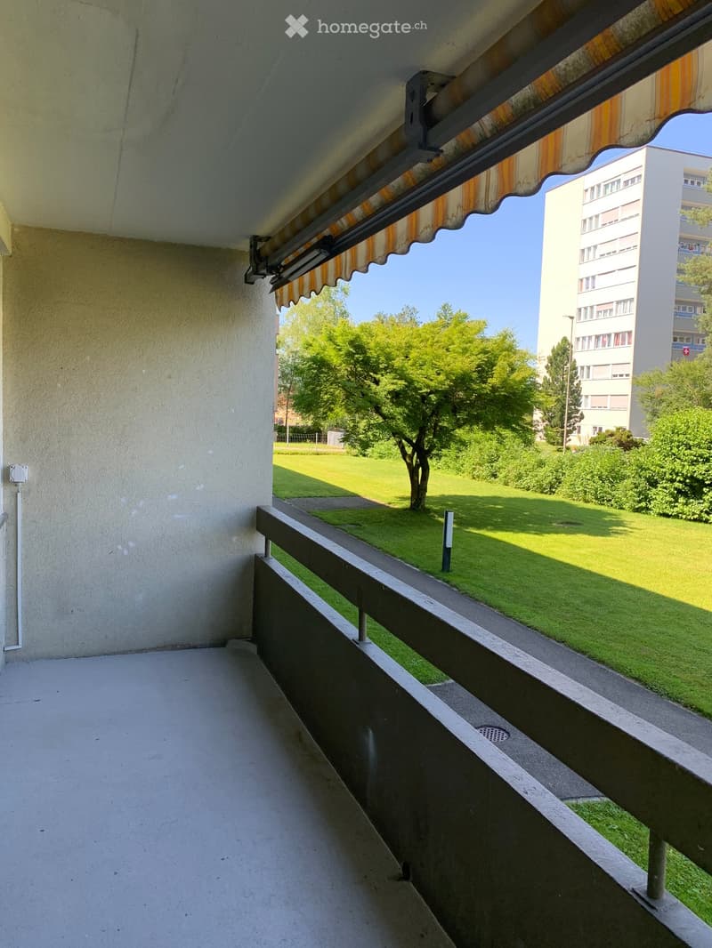 Balcony with a metal railing, striped awning, and view of a green park and residential building.