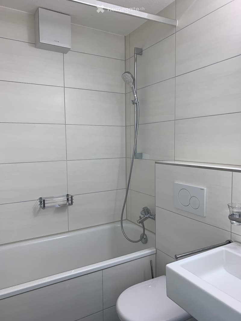 Modern bathroom with white tiles, bathtub, toilet, sink, showerhead, and towel rack.