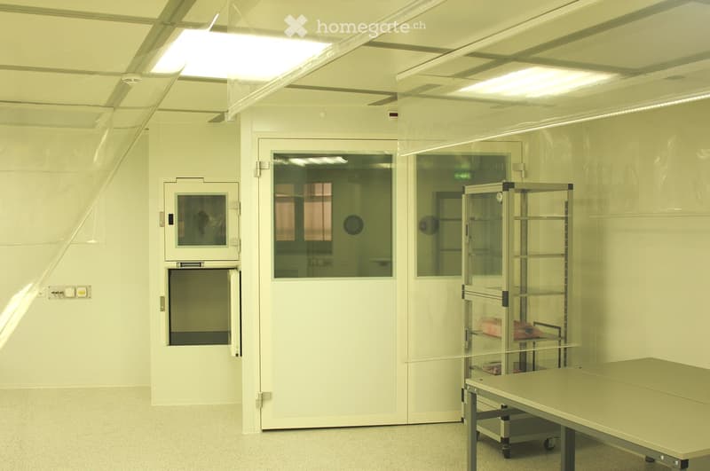 A clean, well-lit room with white walls and a ceiling, featuring a large table, a metal shelving unit with items, and several doors including a large central door with a window and smaller side doors.