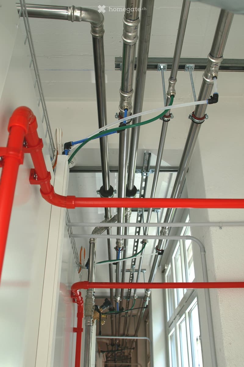 A view of exposed plumbing and electrical systems in a ceiling area, featuring various metal pipes, fittings, and wires.