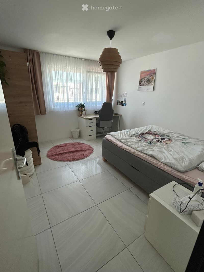 A modern bedroom with a bed, desk, chair, and a hanging lamp. The room features tiled flooring, a round pink rug, and a window with curtains.