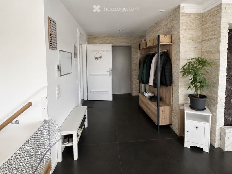 Modern hallway with black tiled floor, white walls, coat rack with jackets, shoe bench, potted plant, and decorative sign.