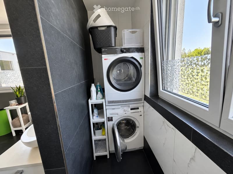 Modern bathroom with a washing machine and dryer stacked, a toilet, a small shelf with cleaning supplies, and a potted plant near the window.