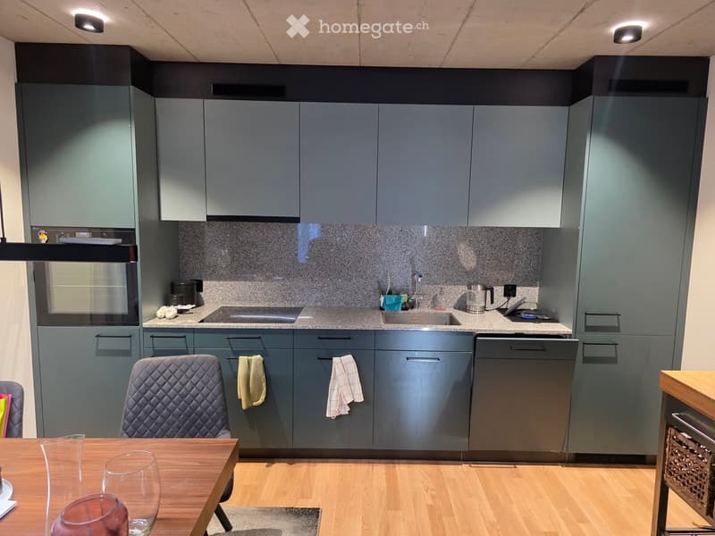 Modern kitchen with grey cabinets, a wooden dining table, and a stainless steel sink. The space includes a dishwasher, oven, and various kitchen items on the counter.