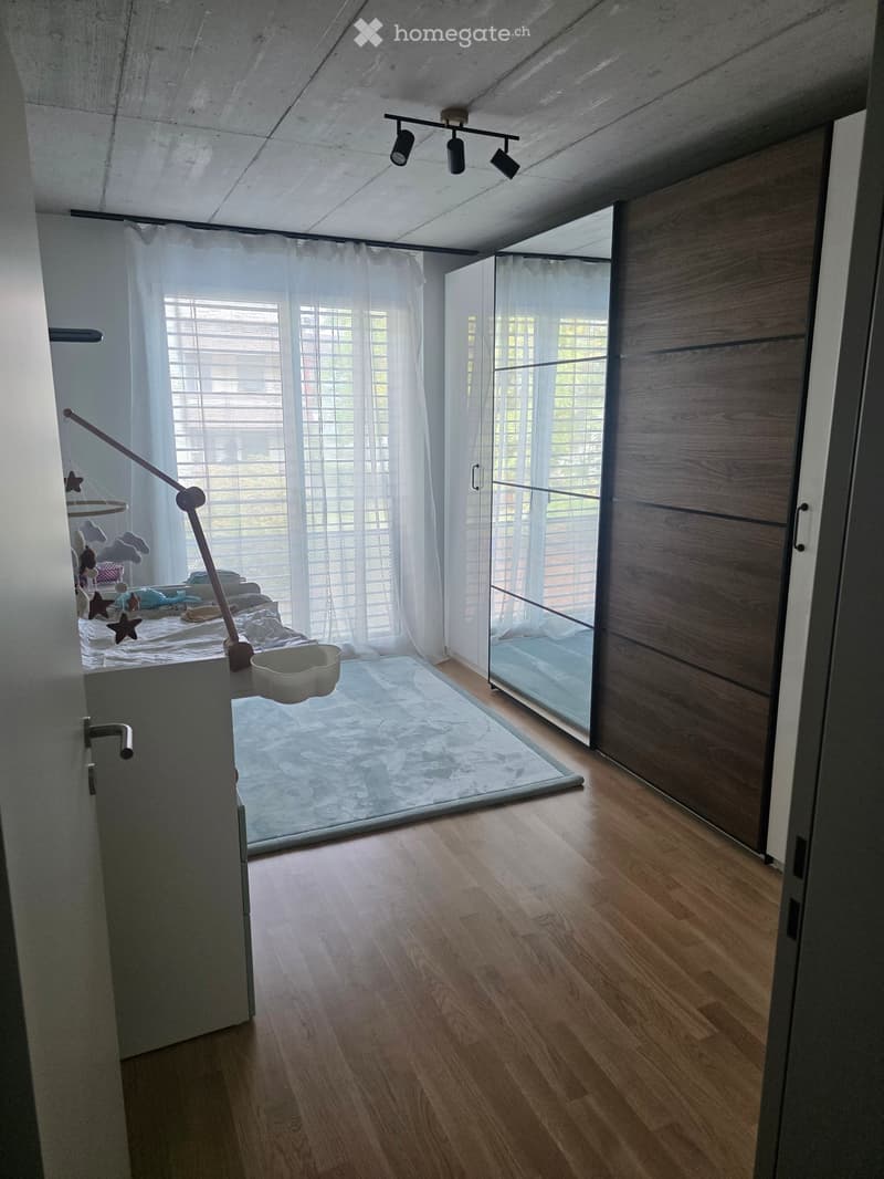 Modern, minimalist bedroom with wooden flooring, large windows with blinds, and a bed with a baby swing and bowl nearby.
