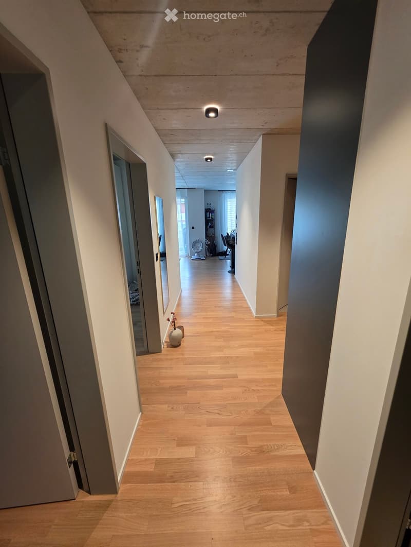 A long, narrow hallway with light wooden flooring, white walls, and recessed ceiling lighting. A small vase with flowers sits on the floor near a partially open door on the left, and another room is visible at the end of the hallway.