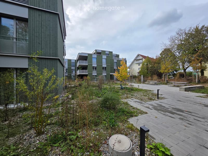 Modern residential complex with green facades, balconies, and a landscaped outdoor area featuring pathways, benches, and small plants.