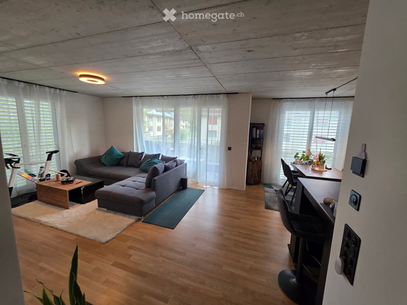 Modern living room with wooden flooring, large windows, grey sectional sofa, coffee table, bar stools, dining table, and a small bookshelf.