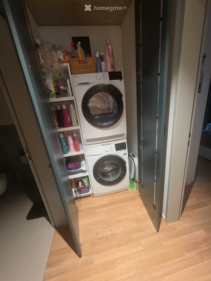 A small bathroom with a washing machine and dishwasher stacked vertically, a shelf with cleaning supplies, and a wooden floor.