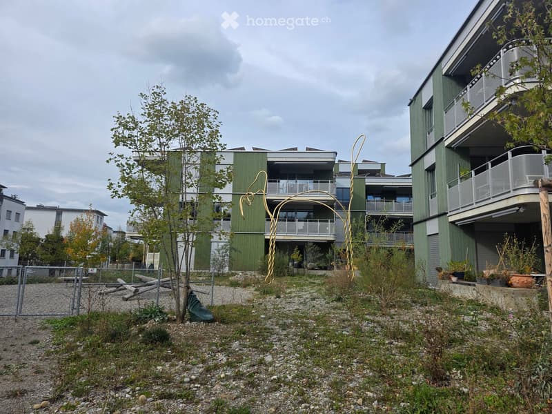 Modern multi-story residential building with green facade, balconies, and surrounding greenery.