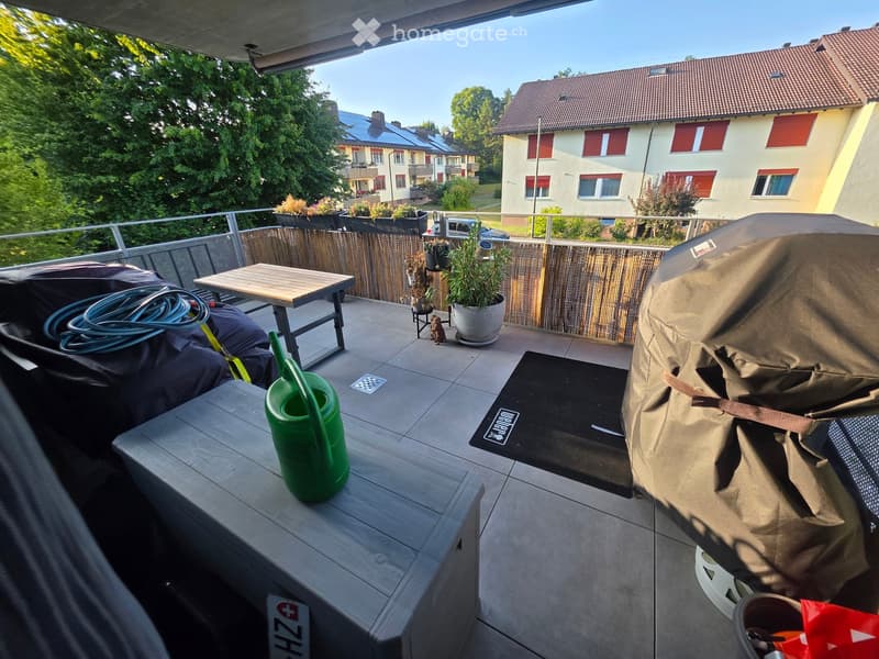 Balcony with a wooden table, chairs, potted plants, a garden hose, a watering can, and a covered grill.