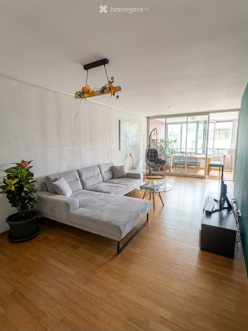 Modern living room with grey sectional sofa, coffee table, TV stand, wicker hanging chair, dining area with table and chairs, and a guitar leaning against the wall.