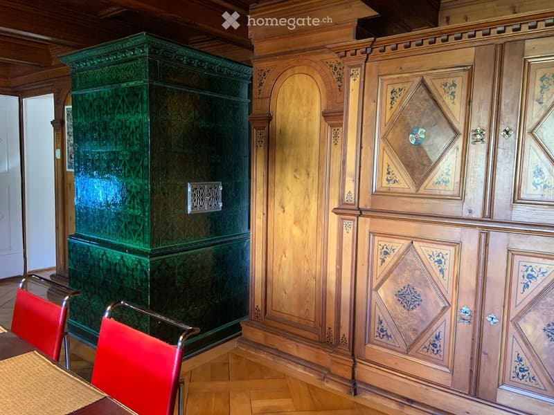 Ornate wooden wall paneling with intricate carvings, green decorative cabinet, red chairs, and a woven table runner.