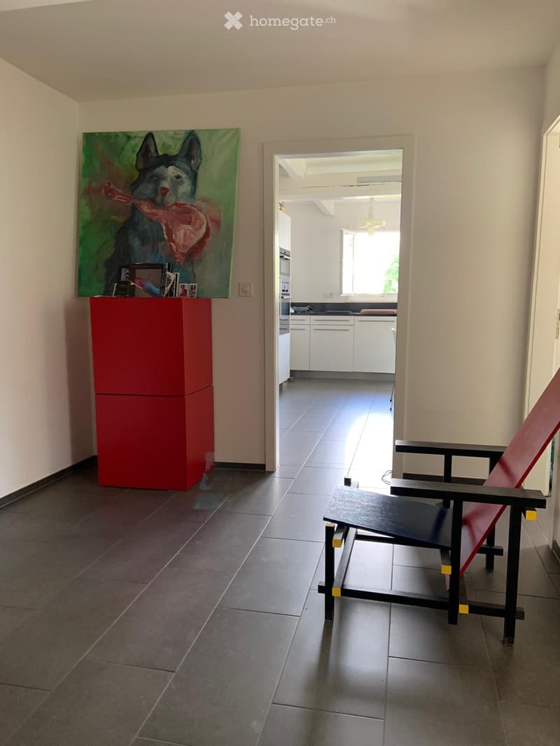 A modern, minimalist hallway with a red chair, a small table, a colorful painting, and a view into a bright kitchen area.