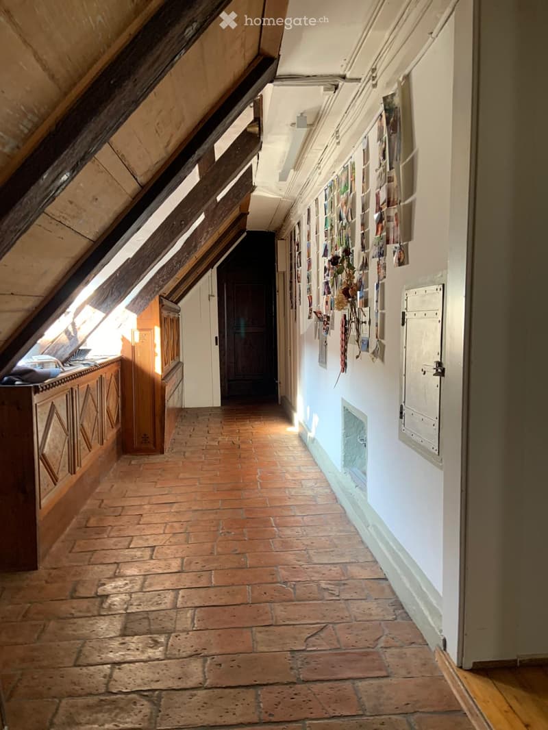 A narrow, slanted attic corridor with exposed wooden beams, brick flooring, and white walls adorned with framed pictures and decorations.