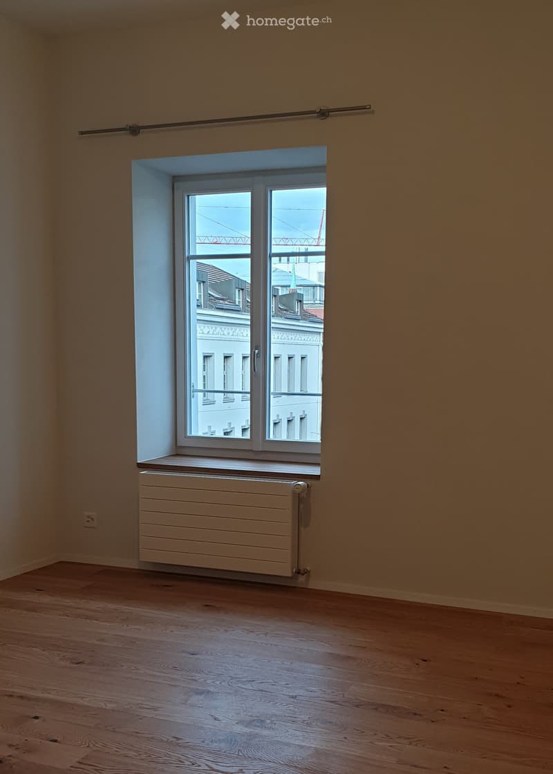 Empty room with wooden flooring, white walls, a window with white frame, radiator, and a curtain rod on the wall.
