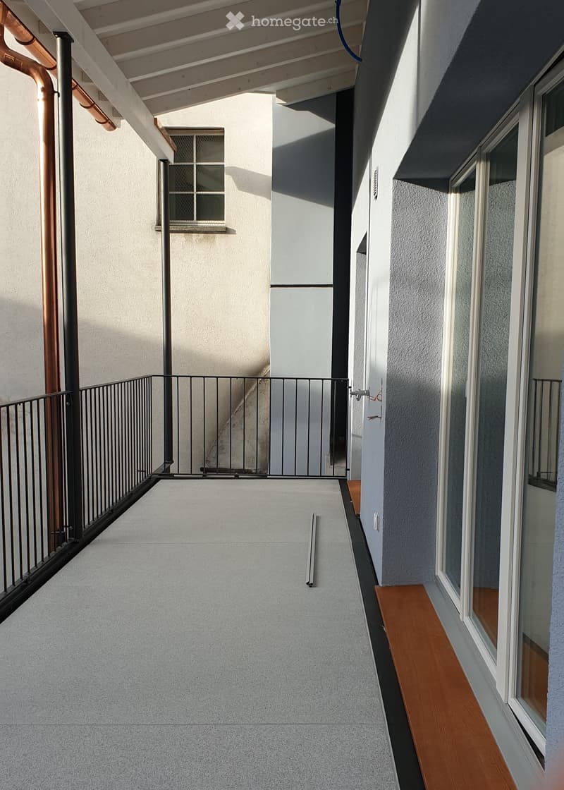 Balcony with grey concrete floor, metal railing, small wooden bench, and windows with white frames.