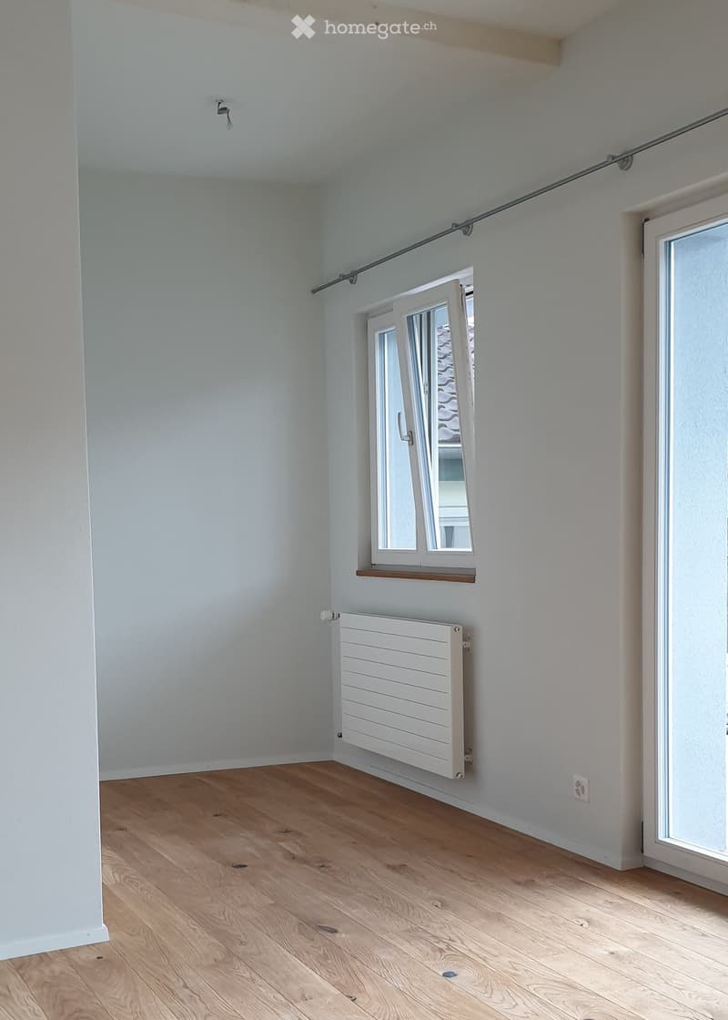 Empty room with light grey walls, wooden floor, white radiator, and large window with sliding doors.