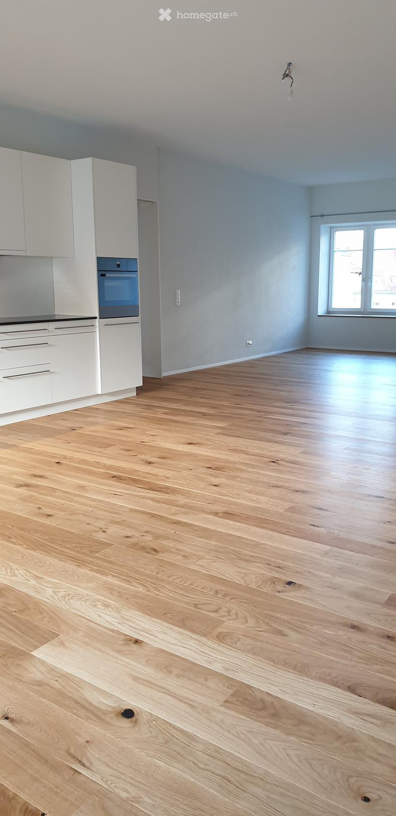 Empty kitchen with light wooden flooring, white cabinets, and a built-in oven. The space is clean and unfurnished.