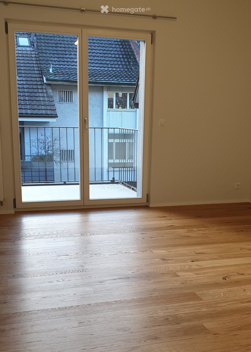 Empty room with wooden flooring and large sliding glass doors leading to a balcony with a view of a neighboring building and roof.