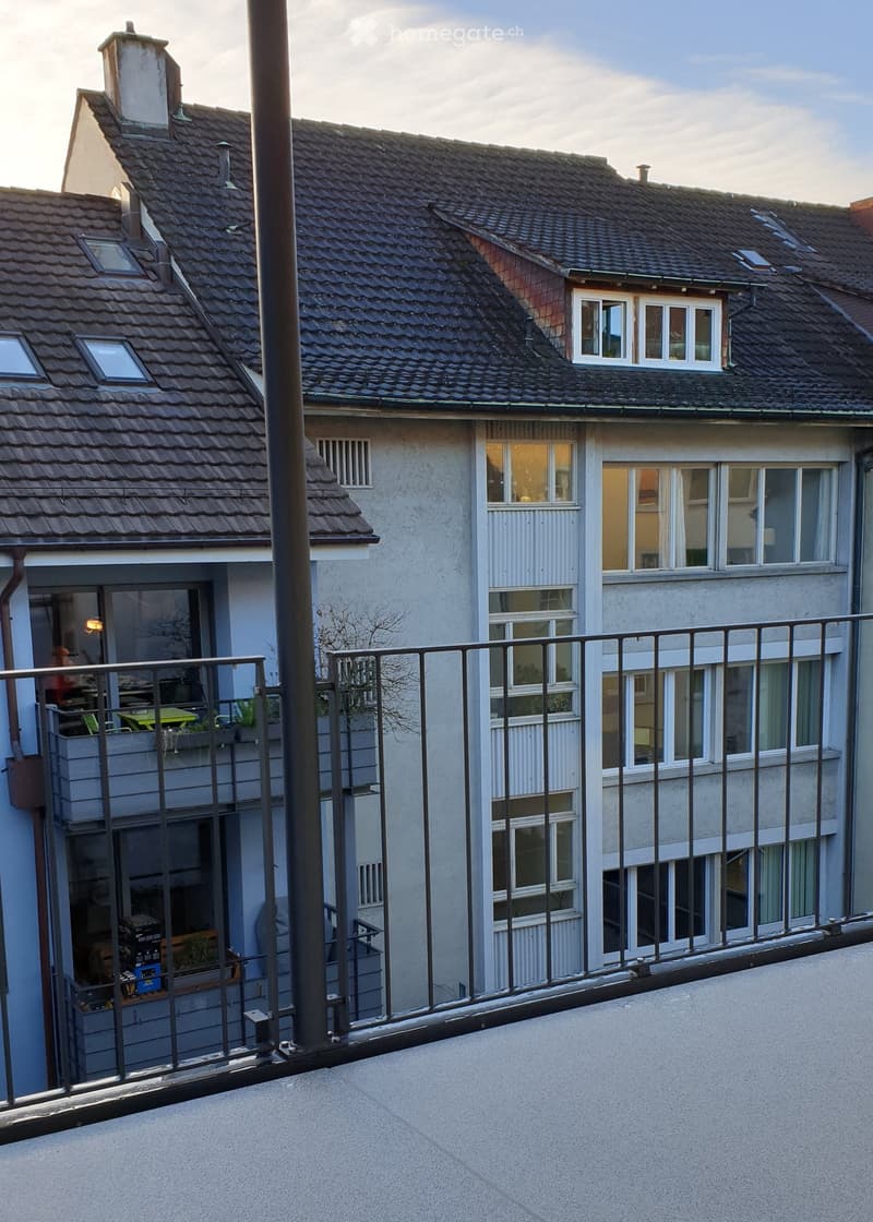 Balcony overlooking a multi-story residential building with tiled roofs and multiple windows. The balcony features a metal railing and a concrete floor.