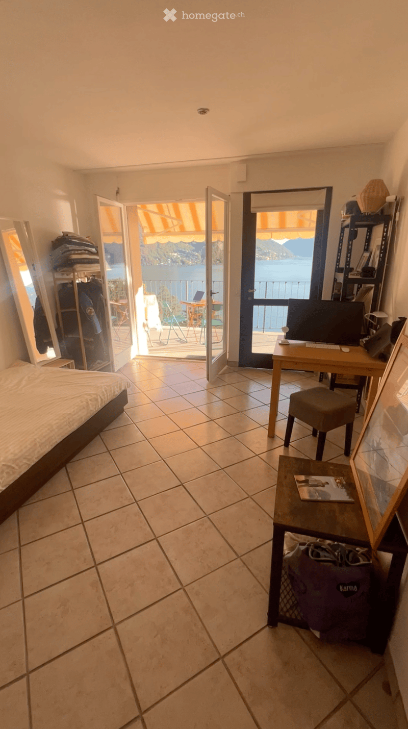 Bright bedroom with tiled floor, bed, desk, and open sliding doors leading to a balcony with a scenic lake view.
