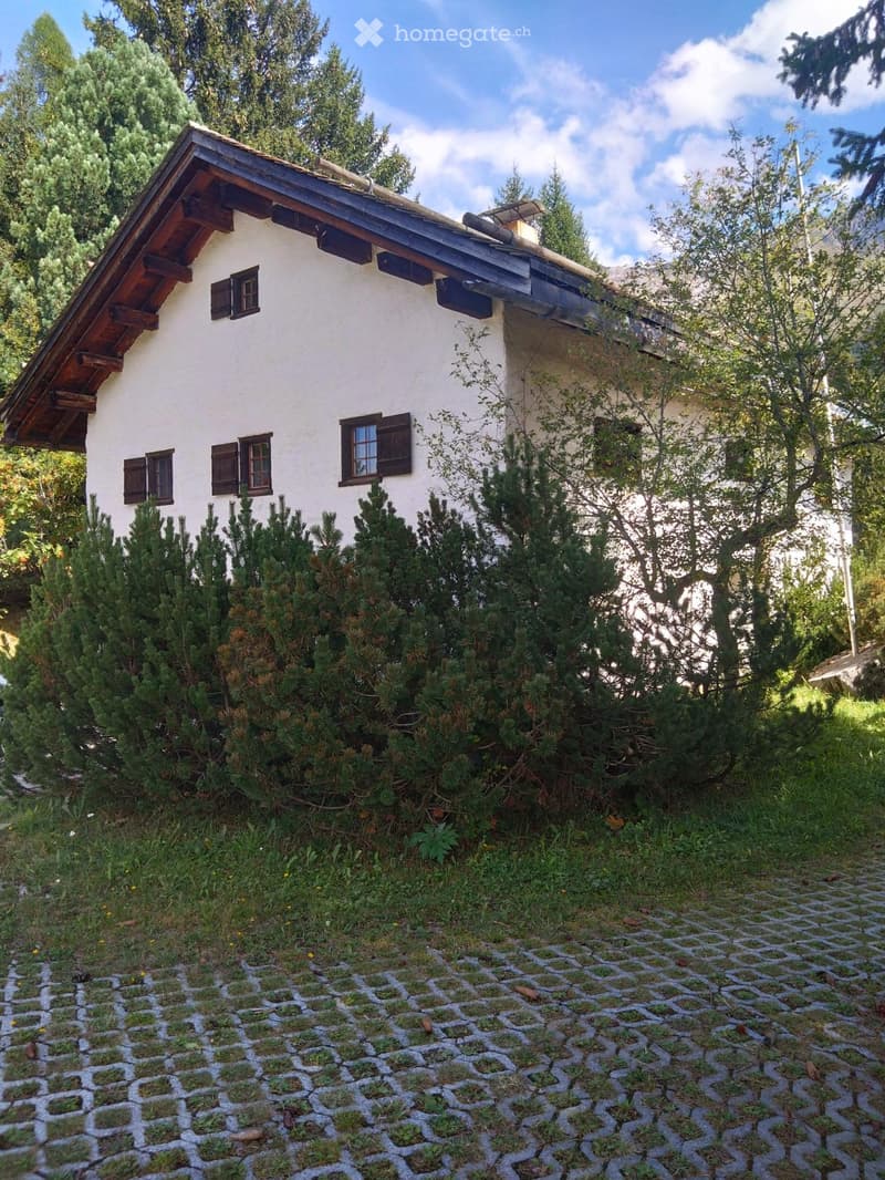 A small, white, single-story house with a wooden roof and dark shutters, surrounded by greenery and a stone-paved pathway.