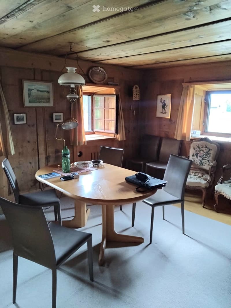 A cozy, wooden-paneled room with a round wooden table surrounded by chairs, a hanging lamp, and various decorative items including framed pictures and a bottle on the table.