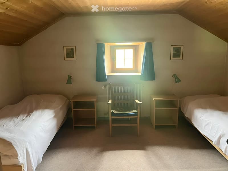 Cozy attic bedroom with two single beds, nightstands, a small chair, and framed pictures on the walls. Natural light streams in through a window with blue curtains.