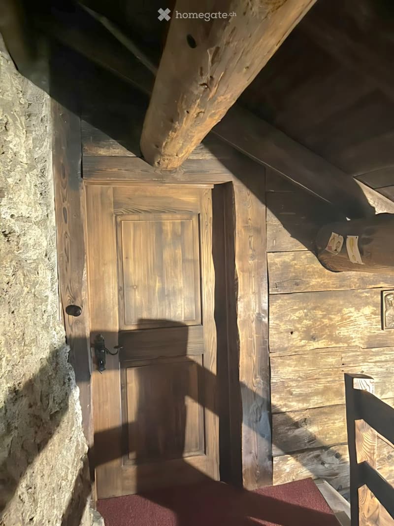 A narrow wooden hallway with exposed wooden beams and a stone wall on one side, featuring a wooden door and a small red rug on the floor.