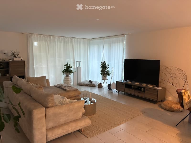 Modern living room with a large sectional sofa, coffee table, TV, side table, and potted plants. The space features large windows with white curtains and a tiled floor.