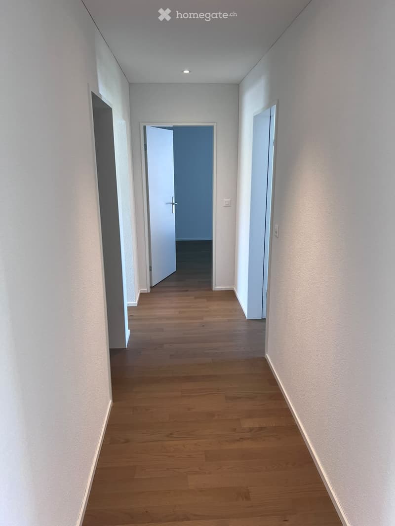 Empty hallway with light wooden flooring, white walls, and two open doors leading to other rooms.