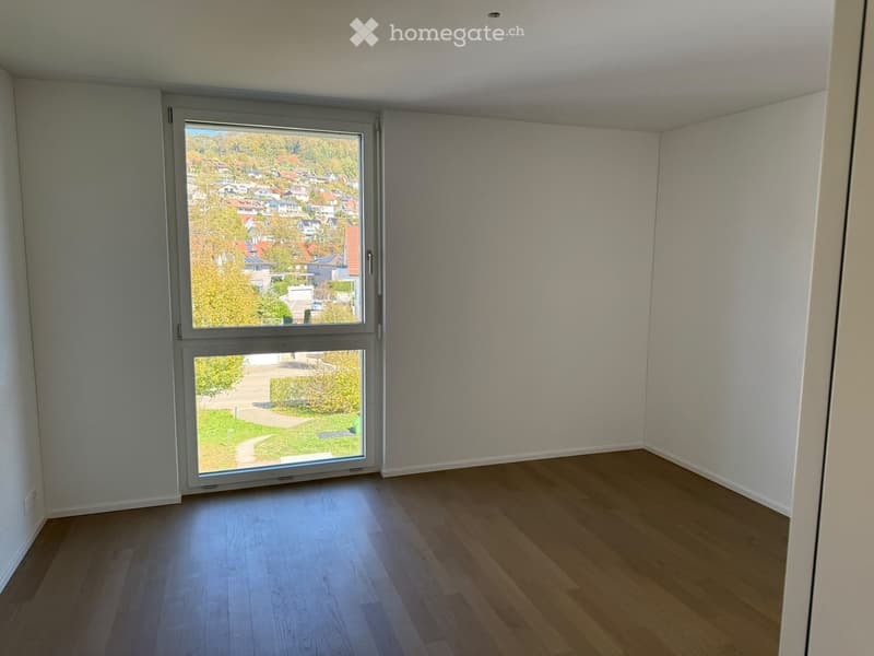 Empty room with wooden flooring and large windows offering a view of a residential area and greenery outside.