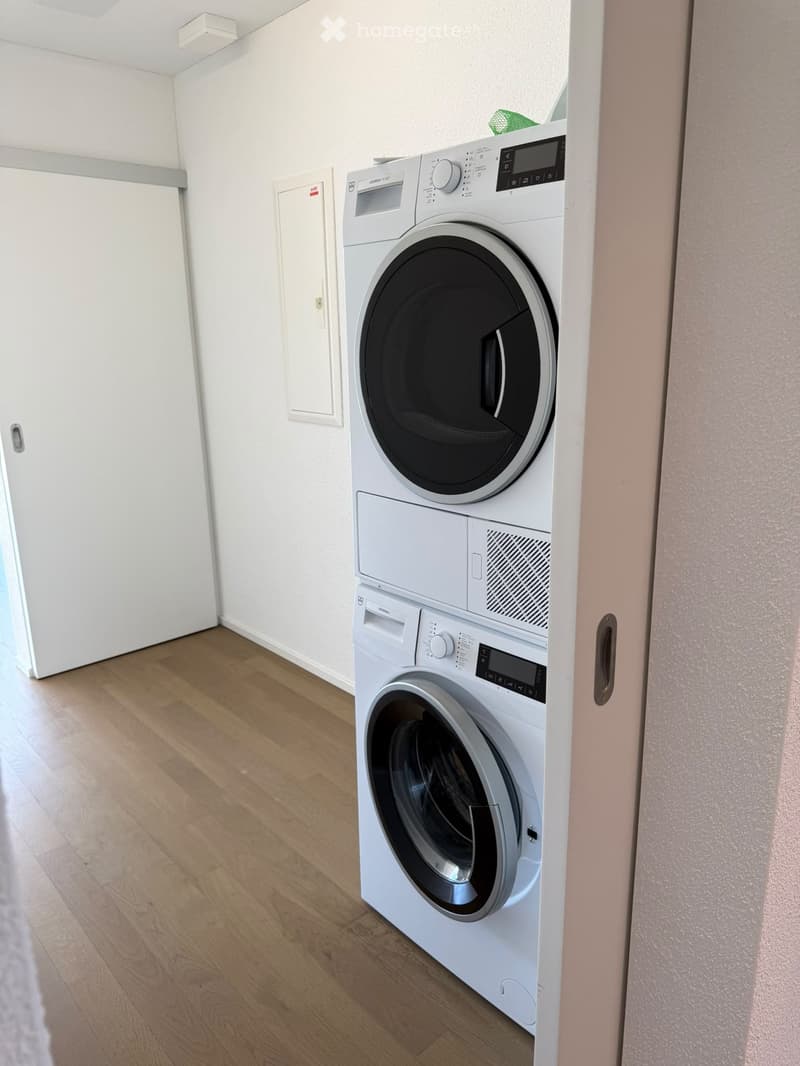 A small, well-lit room with light wooden flooring and white walls, featuring a stacked washing machine and dryer unit.