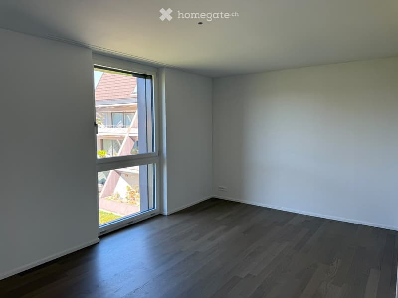 Empty room with light grey walls, wooden flooring, and large window offering view of neighboring building and garden.