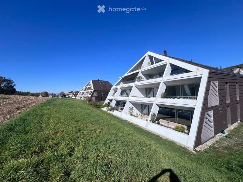 Modern, multi-level house with white and brown exterior, large windows, and balconies, situated on a grassy slope with surrounding greenery.