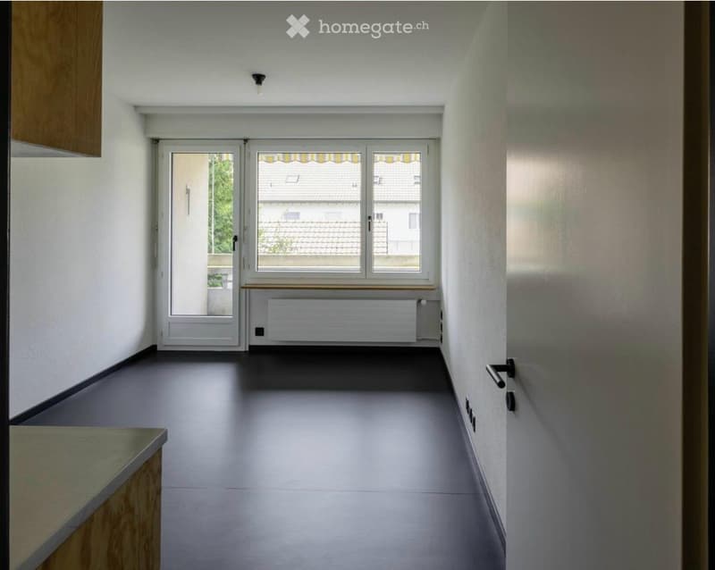 Empty hallway with grey flooring, white walls, a window with blinds, and a radiator below it. The space is clean and unfurnished.
