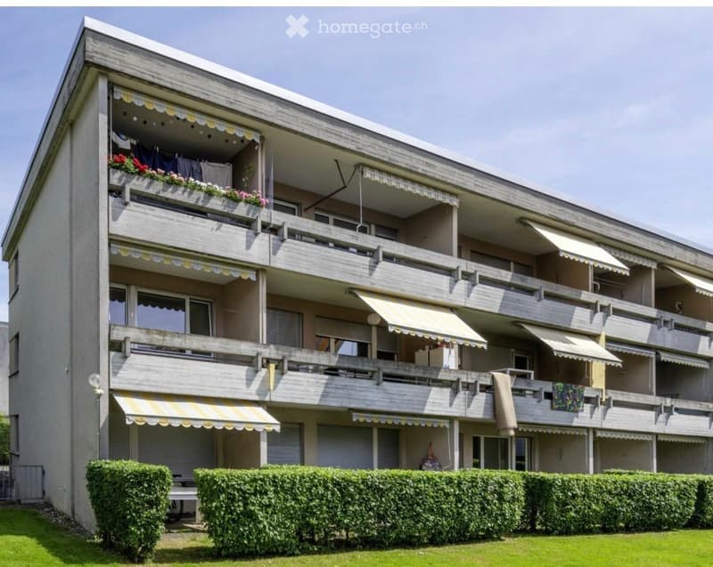 Multi-story apartment building with balconies, awnings, and flower boxes. Green hedges and lawn in front.