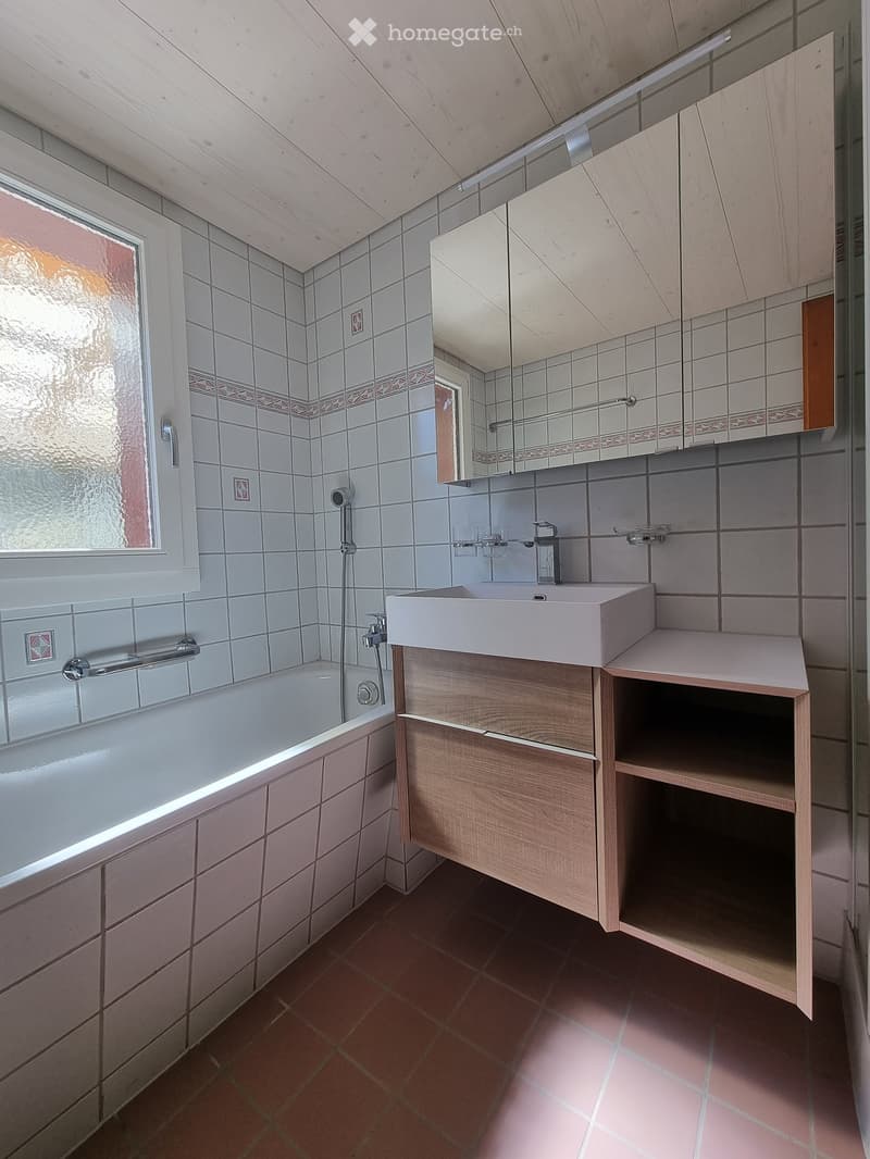 Modern bathroom with a bathtub, sink, mirror, and wooden cabinet. The room features tiled walls and a window with frosted glass.