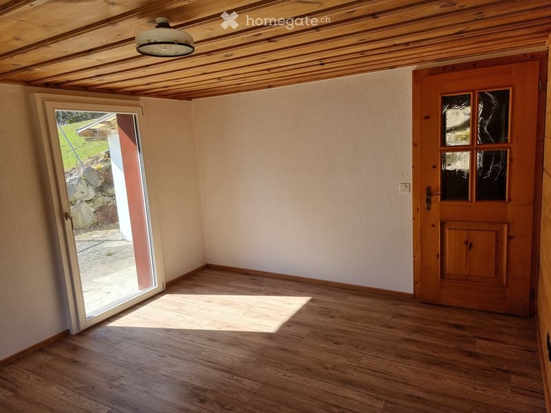 Empty room with wooden flooring, exposed wooden ceiling beams, and a large open door leading outside. The room has white walls and a pendant light fixture.