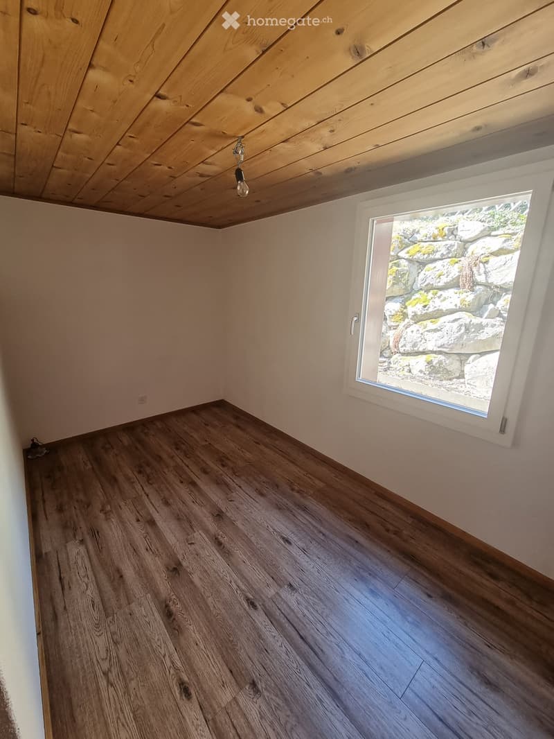 Empty room with wooden flooring, white walls, and a large window showing a view of rocks and greenery outside.