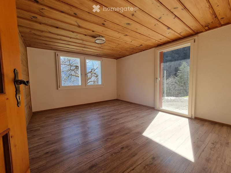 Empty room with wooden flooring, exposed wooden ceiling beams, and large windows with a view of trees. A sliding glass door leads to an outdoor area.