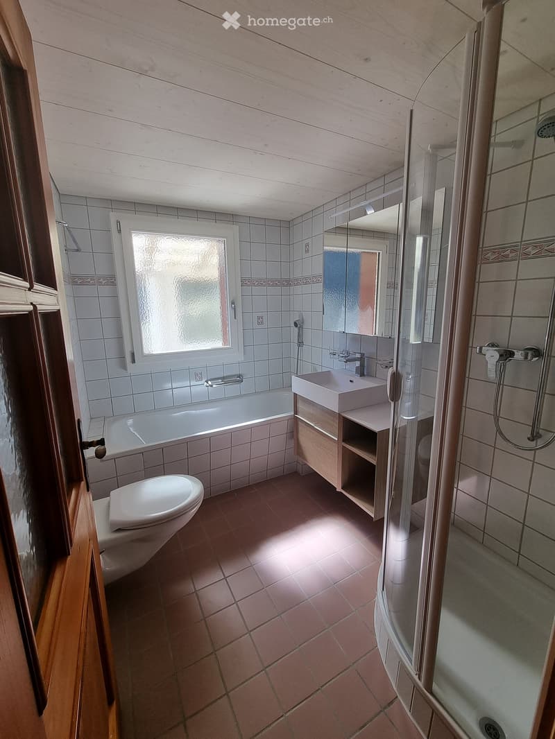 Bathroom with a bathtub, toilet, sink, mirror, and a glass-enclosed shower. The room features tiled walls and a window with frosted glass.