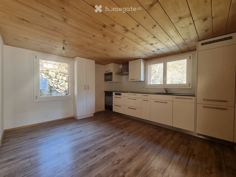 Modern kitchen with light-colored cabinets, wooden flooring, and large windows offering a view of a stone wall outside. Features include a sink, oven, microwave, and ample cabinet space.