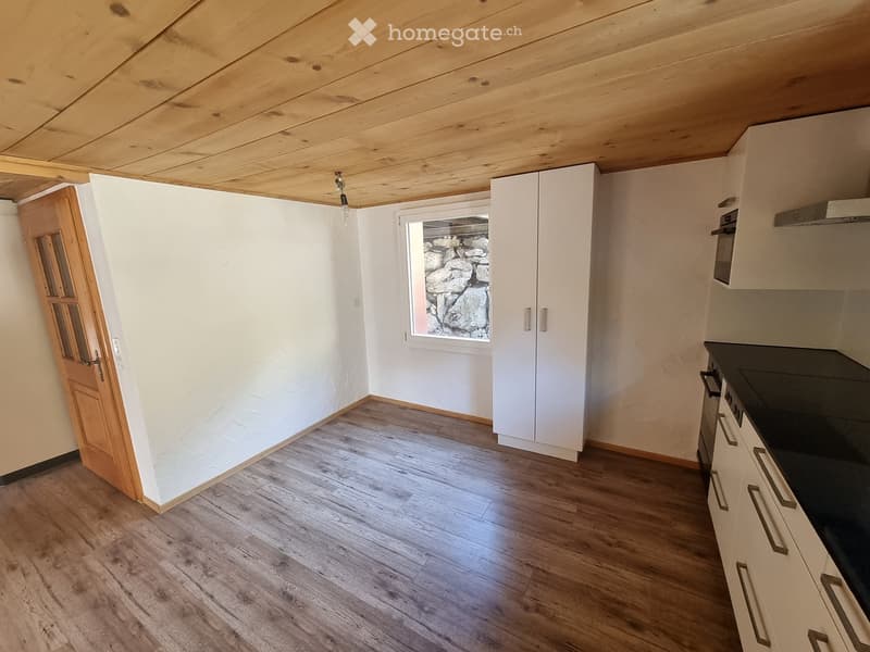 Empty kitchen with wooden flooring, white cabinets, and a window overlooking a stone wall exterior.