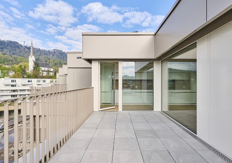 Modern balcony with large glass doors, tiled floor, and a view of a town with a church and mountains in the background.