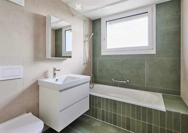 Modern bathroom with a white sink, mirror, bathtub, and toilet. Green tiles line the walls and floor, and a window provides natural light.