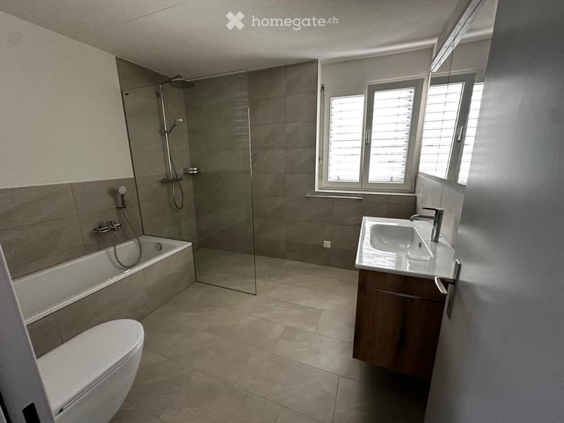 Modern bathroom with a bathtub, toilet, sink, and glass-enclosed shower. Features tiled walls and floor, and has three windows with blinds.